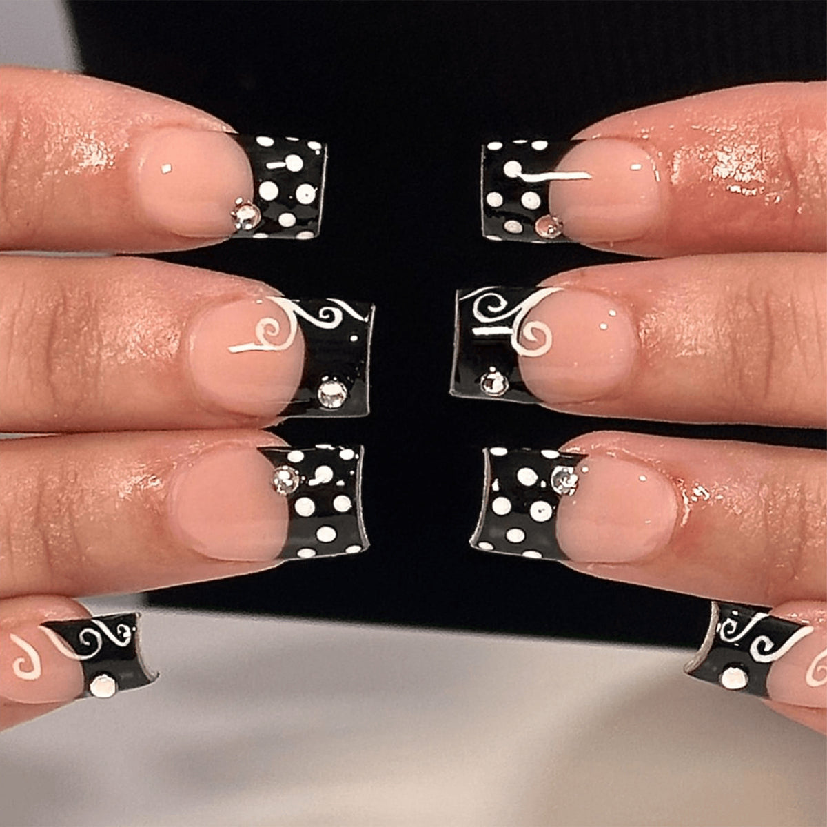 Hands wearing short square press-on nails with black french tips, white swirls and polka dots