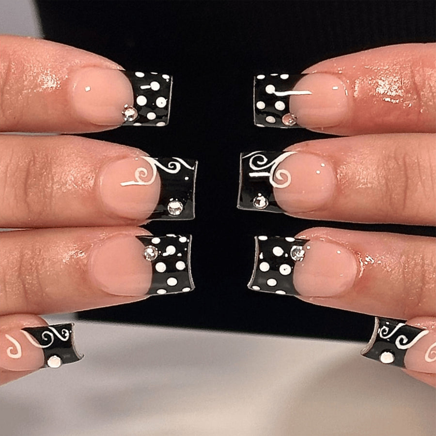 Hands wearing short square press-on nails with black french tips, white swirls and polka dots