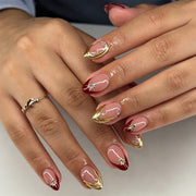 Hands wearing glossy nude and burgundy almond press-on nails with gold swirls and starburst charms.