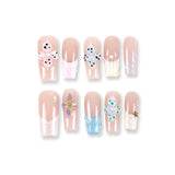 Uñas postizas 365Nails color nude translúcido estilo francés con flores 3D en tonos pastel y detalles de perlas.