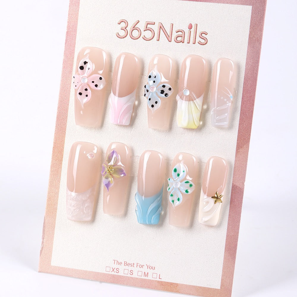 Uñas postizas 365Nails color nude translúcido estilo francés con flores 3D en tonos pastel y detalles de perlas.