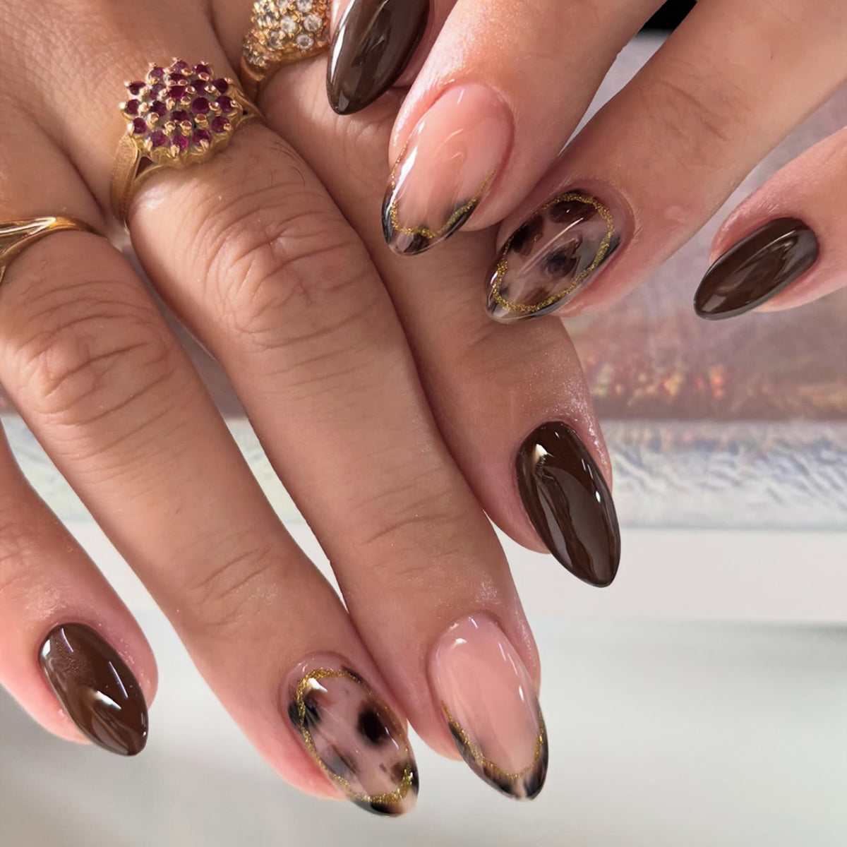 Almond-shaped press-on nails worn on fingers featuring dark brown glossy finish, sheer nude base with leopard spots, and