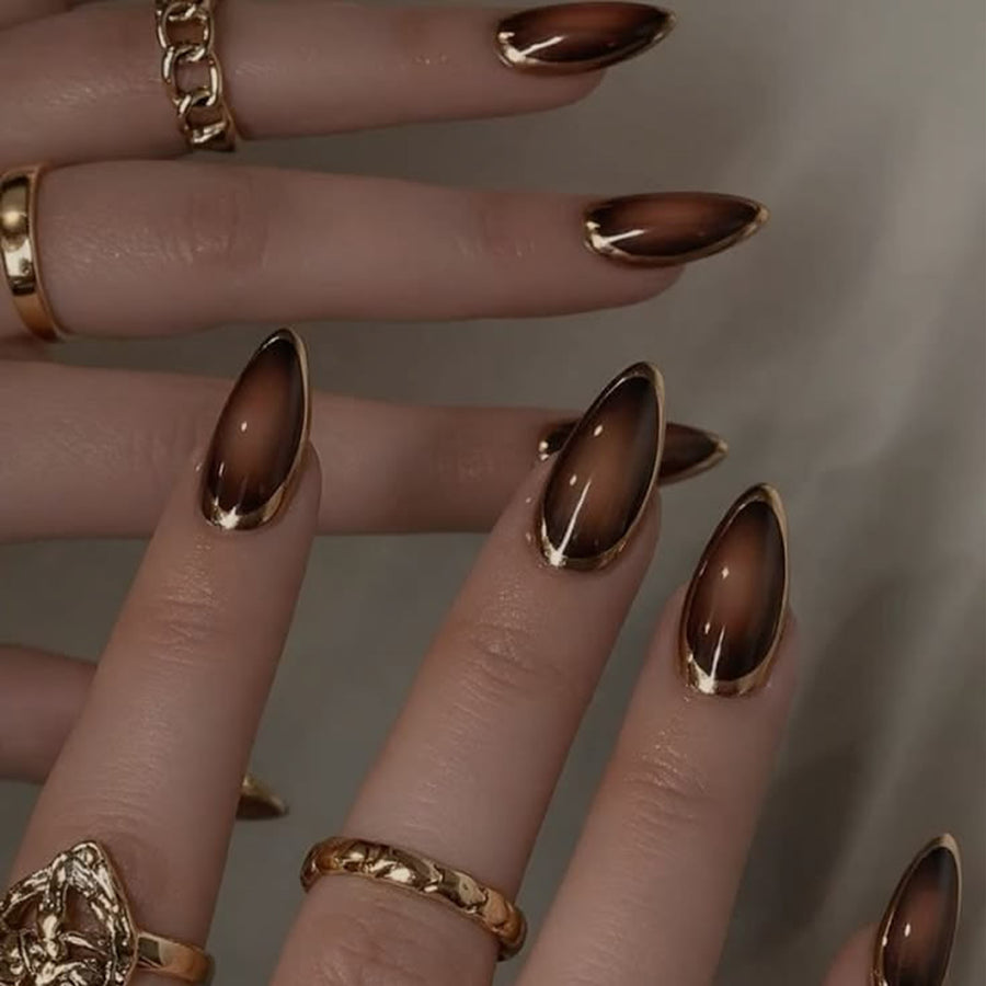 Hand wearing glossy brown gradient almond press-on nails with gold rings.