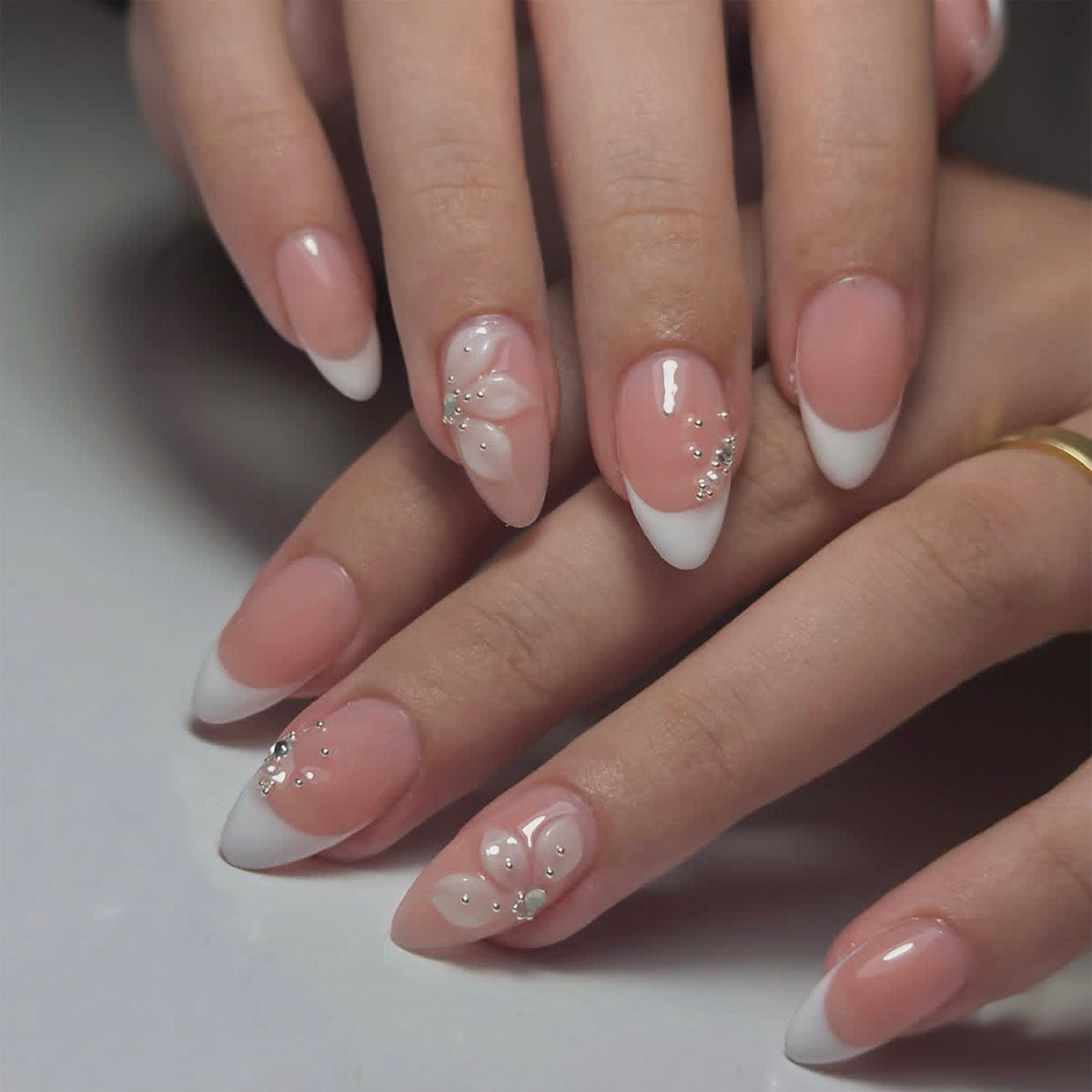 Hand wearing nude French almond press-on nails with white tips, 3D flowers and rhinestones.