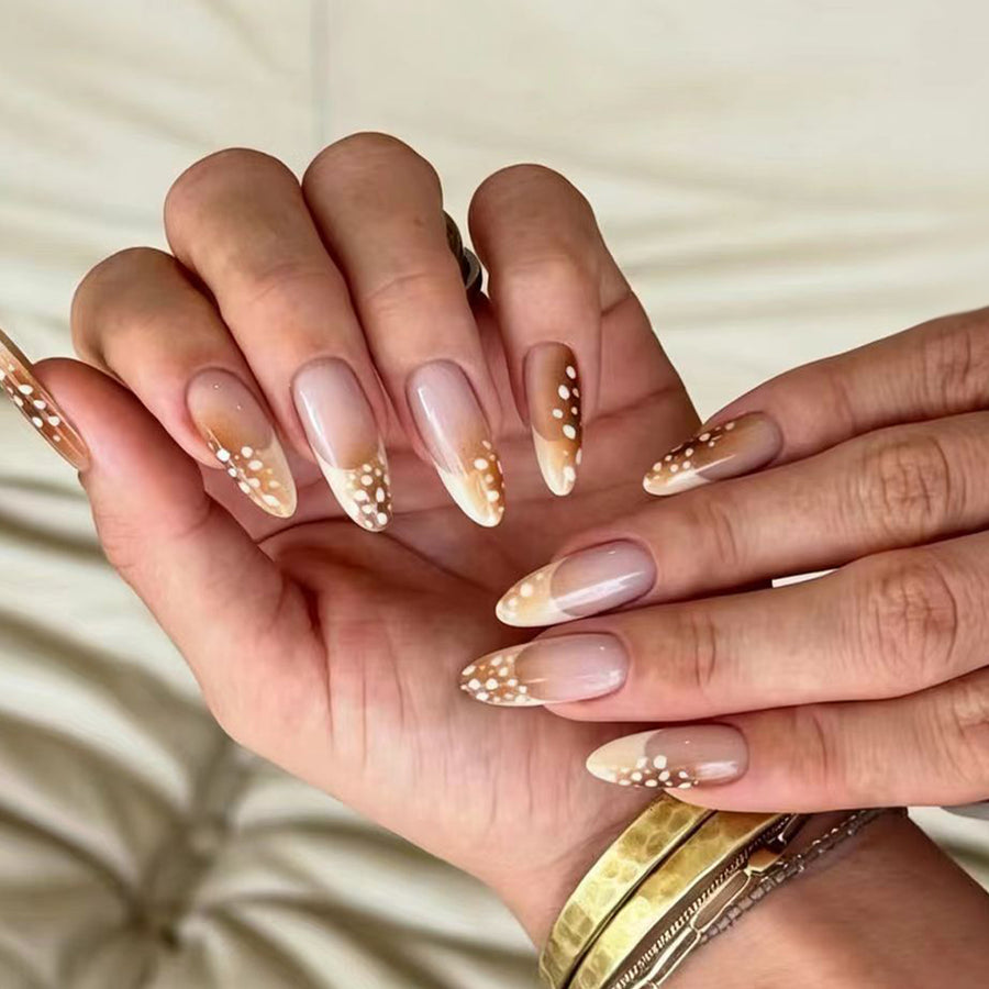 Hand wearing nude almond press-on nails with brown gradient tips and white flecks