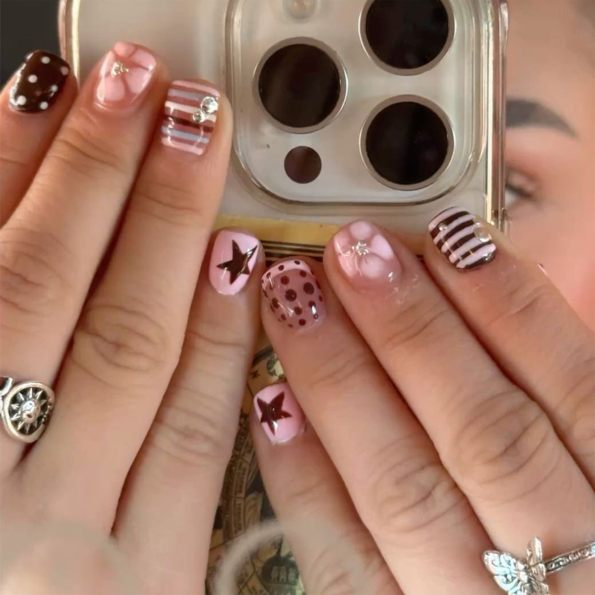 Hand wearing pink and brown press-on nails with stars, dots, and plaid patterns while holding a phone.