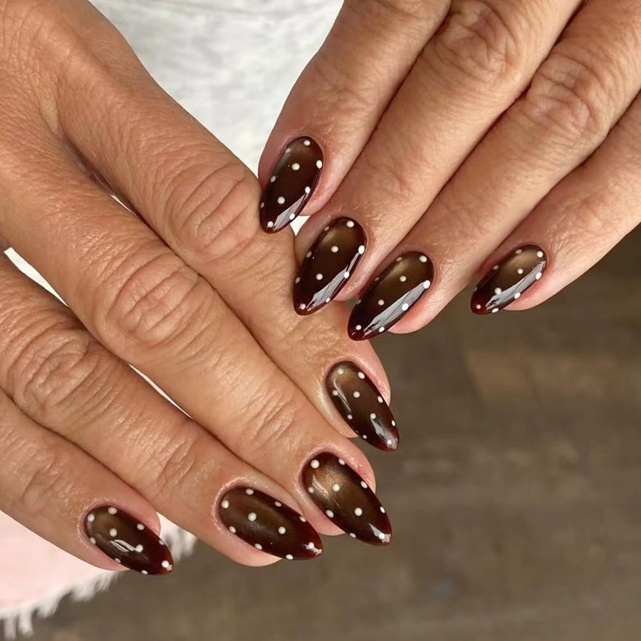 Hands with glossy dark brown almond press-on nails decorated with small white polka dots