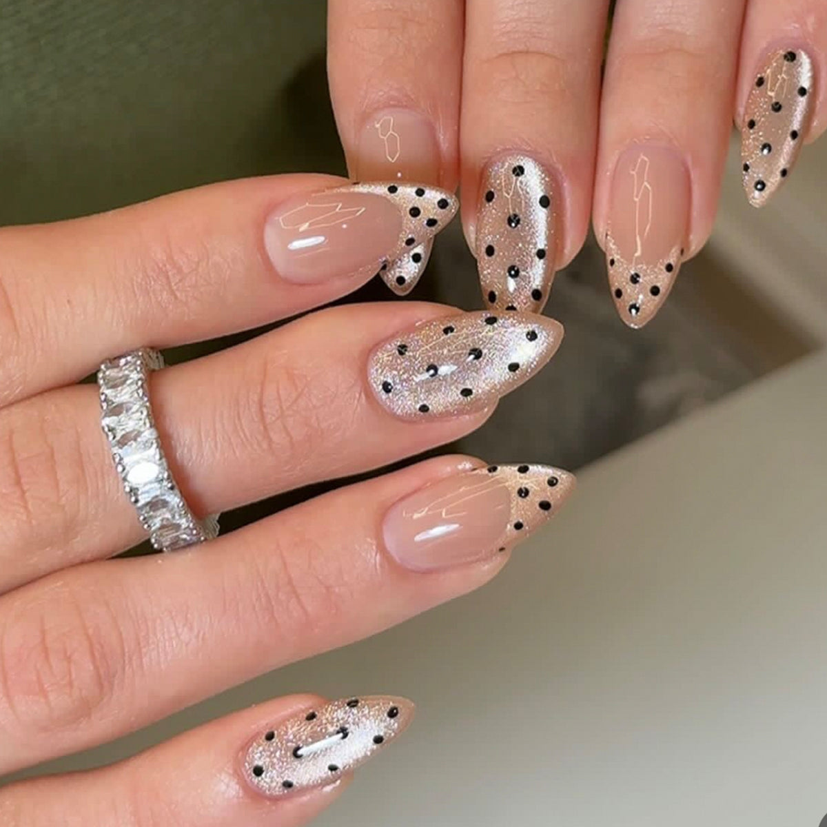 Hand wearing almond-shaped nude press-on nails with black polka dots and fine shimmer